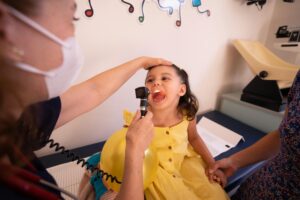 young happy girl in a yellow dress sitting in the doctors office exam room with a female doctor looking into her throat.